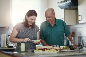 A caregiver providing 24-hour care for an older man prepares a sandwich as he looks on with a smile.