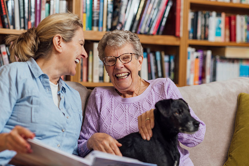 A Sacramento caregiver laughs with an older woman holding her dog as they look at a photo album together.